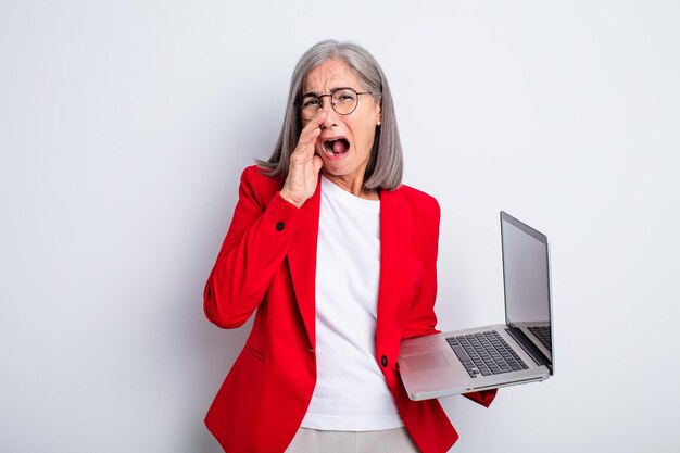 Ältere hübsche Frau, die sich glücklich fühlt und mit den Händen neben dem Mund einen großen Schrei ausspricht. Business- und Laptop-Konzept