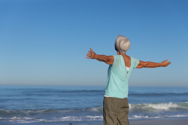 Ältere kaukasische Frau genießt die Zeit am Strand an einem sonnigen Tag, steht mit ausgestreckten Armen und blickt auf das Meer, mit blauem Himmel im Hintergrund