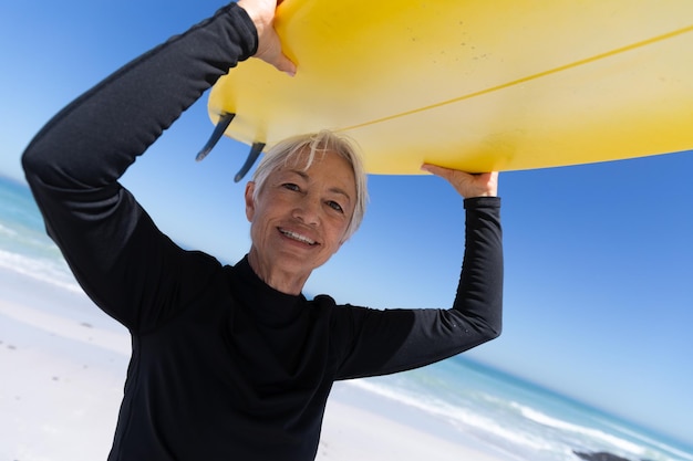 Ältere kaukasische Frau genießt die Zeit am Strand, steht und hält ein Surfbrett über ihrem Kopf