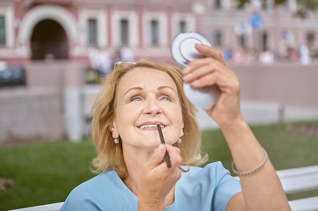 Ältere kaukasische weiße attraktive Dame ungefähr 60 Jahre alt geht in der Innenstadt spazieren und repariert Make-up mit einem Taschenspiegel.