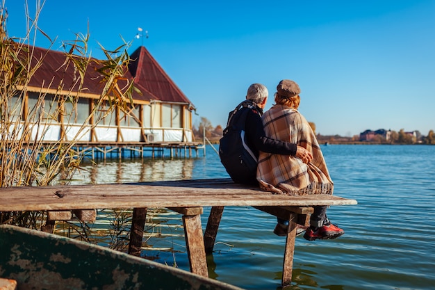 Ältere Paare von den Touristen, die durch Herbstsee sich entspannen. Glücklicher Mann und Frau, die Natur genießt und das Sitzen auf Pier umarmt