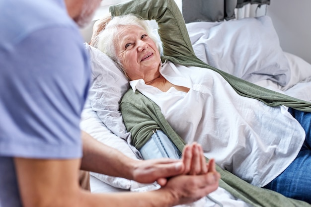 Ältere Patientin im Krankenhaus mit besorgtem Ehemann, der Hände hält, während Blutdruck mit Tonometer prüft. Mann hilft, unterstützt