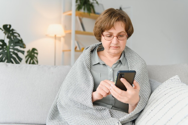 Ältere reife erwachsene Frau mit Smartphone, die zu Hause auf der Couch sitzt Fröhliche Großmutter mit Brille, die mit ihrem Handy im Internet surft Technologiekonzept