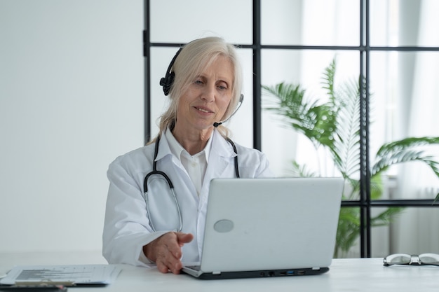 Ältere Ärztin mit Stethoskop, die einen Laborkittel und ein Headset trägt, kommuniziert per Videoanruf mit einem Patienten über einen Laptop