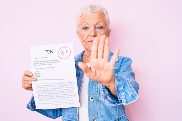 Ältere schöne Frau mit blauen Augen und grauen Haaren zeigt eine bestandene Prüfung mit offener Hand und macht ein Stoppschild mit ernster und selbstbewusster Verteidigungsgeste