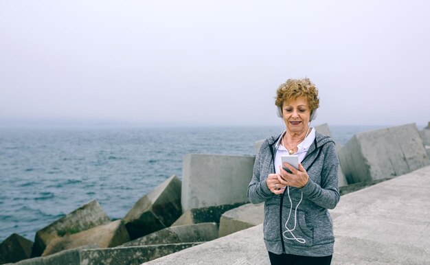 Ältere Sportlerin mit Kopfhörern, die am Seepier auf ihr Smartphone schaut