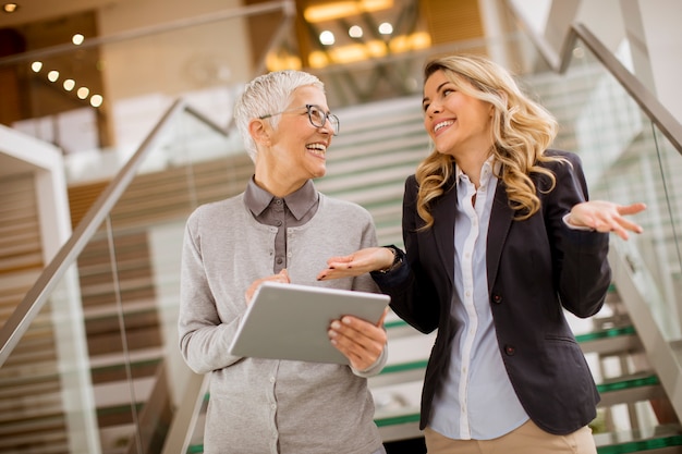 Ältere und junge Geschäftsfrauen, die im Büro mit digitaler Tablette stehen und sich besprechen