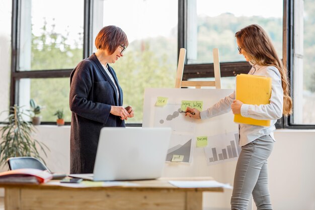 Ältere und junge Geschäftsfrauen unterhalten sich in der Nähe des Vorstands mit Finanzdiagrammen im Büro am Fenster