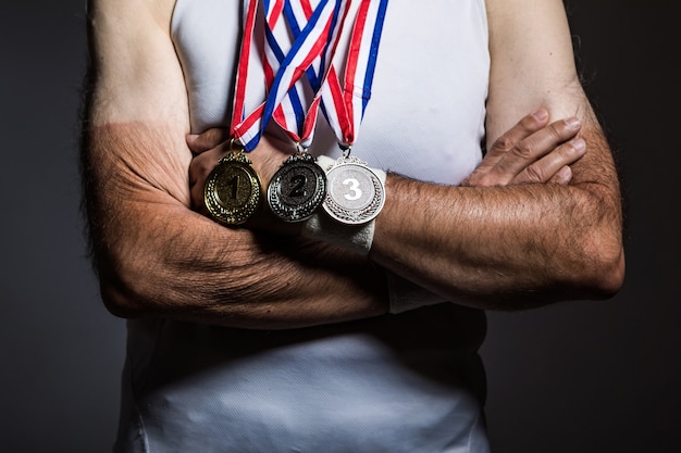 Älterer Athlet mit weißem ärmellosem Hemd, mit Sonnenflecken an den Armen, mit drei Medaillen am Hals, mit verschränkten Armen, auf dunklem Hintergrund. Sport- und Siegkonzept.