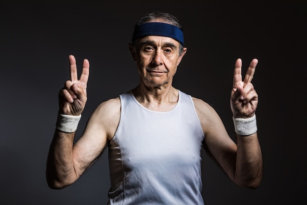 Älterer Athlet mit weißem ärmellosem Hemd mit Sonnenflecken an den Armen und blauem Stirnband, das mit den Fingern das Victory-Zeichen auf dunklem Hintergrund macht. Sport- und Siegkonzept.