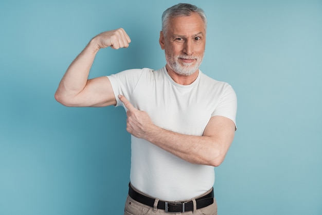 Älterer bärtiger Mann, der Muskeln zeigt, die weißes T-Shirt aufwerfen. Ernsthafter Mann zeigt auf seinen perfekten Bizeps