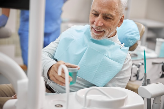 Älterer Bürger, der auf einem Zahnarztstuhl sitzt, bekommt eine Tasse Wasser, um sich während der zahnärztlichen Behandlung den Mund auszuspülen
