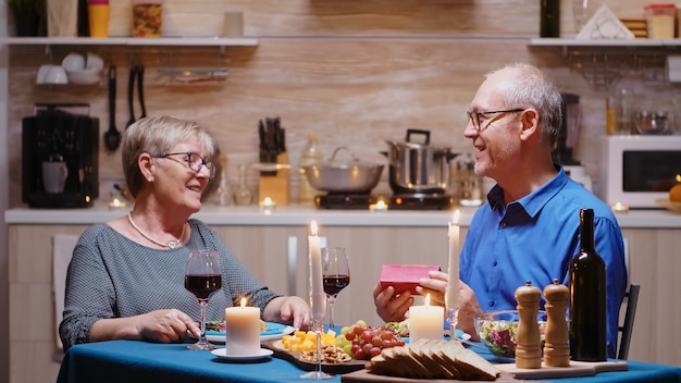 Älterer Ehemann, der der älteren Frau beim romantischen Abendessen in der Küche ein Geschenk gibt. Fröhliches, fröhliches älteres Ehepaar, das zu Hause zusammen isst, das Essen genießt, ihr Jubiläum feiert, Überraschungsholi