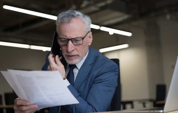 Älterer Geschäftsmann, der auf Handy spricht, Vertrag liest, Brainstorming arbeitet im Büro. Multitasking-Konzept