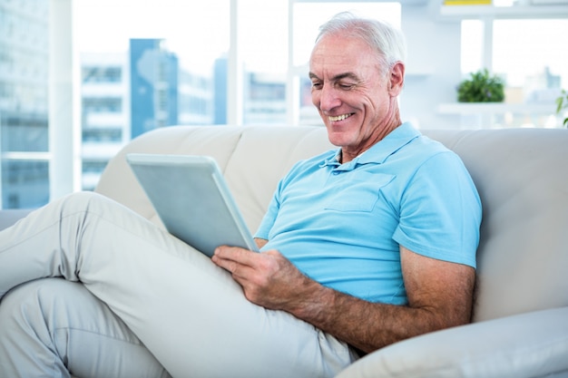 Älterer glücklicher Mann, der auf Sofa bei der digitalen Tablette zu Hause sitzt