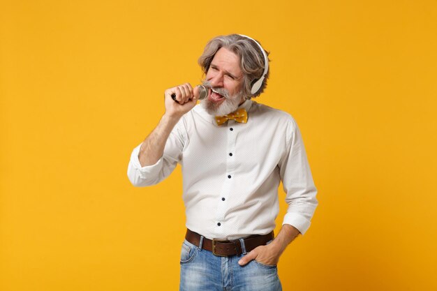 Älterer grauhaariger bärtiger Mann mit Schnurrbart in weißer Hemdfliege isoliert auf gelbem Wandhintergrund. People-Lifestyle-Konzept. Mock-up-Kopienbereich. Hören Sie Musik mit Kopfhörern, singen Sie ein Lied im Mikrofon.