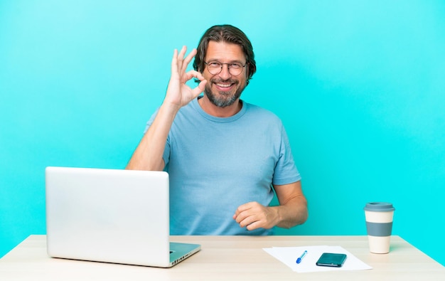 Älterer holländischer Mann in einem Tisch mit einem Laptop isoliert auf blauem Hintergrund, der mit den Fingern ein Ok-Zeichen zeigt