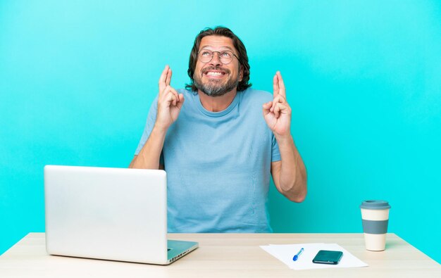 Älterer holländischer Mann in einem Tisch mit einem Laptop isoliert auf blauem Hintergrund mit gekreuzten Fingern