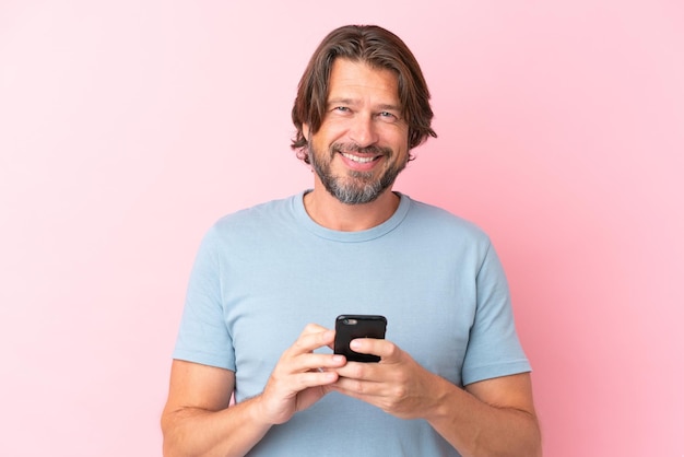 Älterer holländischer Mann isoliert auf rosa Hintergrund, der in die Kamera schaut und lächelt, während er das Handy benutzt