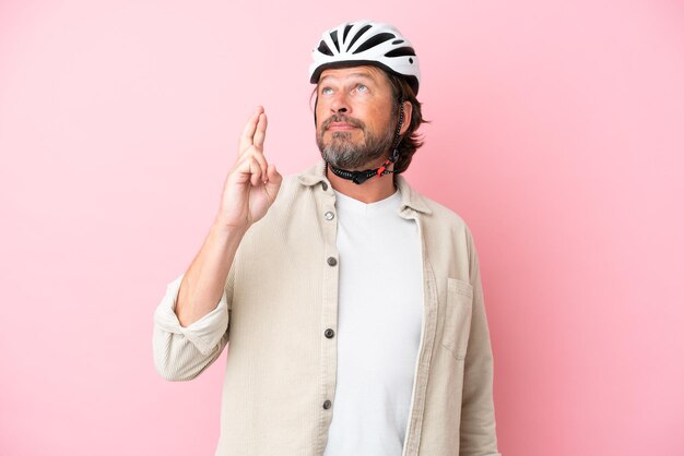 Älterer holländischer Mann mit Fahrradhelm isoliert auf rosa Hintergrund mit gekreuzten Fingern und wünscht das Beste