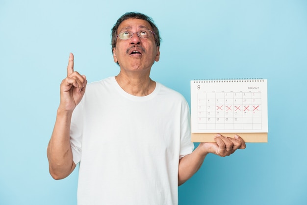 Älterer indischer Mann, der einen Kalender lokalisiert auf blauem Hintergrund hält, der mit geöffnetem Mund nach oben zeigt.