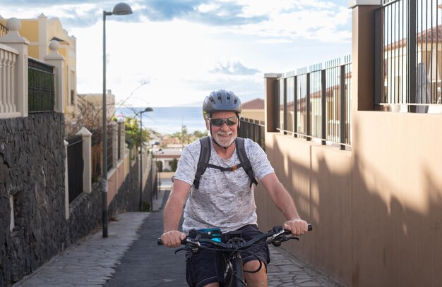 Älterer lächelnder Mann mit Sporthelm und Sonnenbrille, der bei Sonnenuntergang mit seinem Elektrofahrrad in bergauf gehender Gasse radelt. Horizont über Wasser im Hintergrund