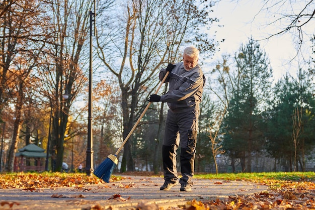 Älterer männlicher Arbeiter fegt trockene Blätter mit Besen im Freien, Blick von unten auf einen grauhaarigen Mann