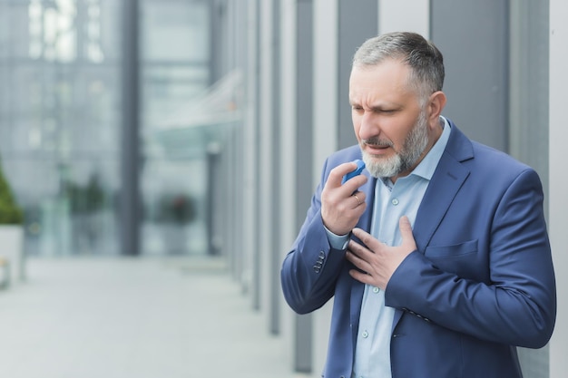 Älterer männlicher Geschäftsmann, der in der Nähe des Bürochefs krank ist, verwendet einen Inhalator, um das Atmen des grauhaarigen Bankiers zu erleichtern