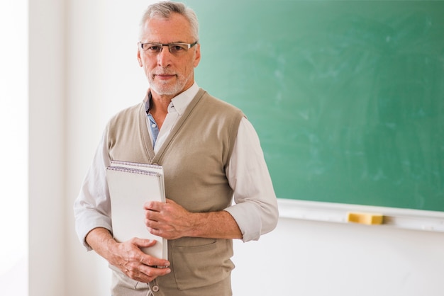 Älterer männlicher Professor in den Gläsern, die das Notizbuch steht gegen Tafel halten