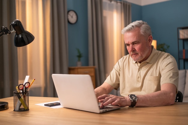 Älterer Mann, der abends mit Laptop am Schreibtisch sitzt, arbeitet in einem Salon