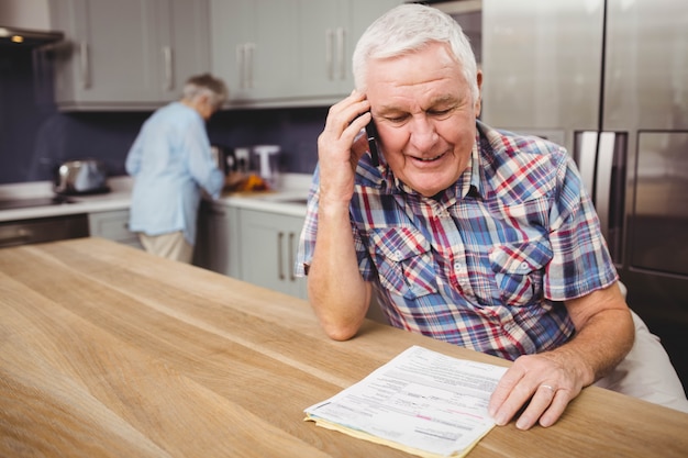 Älterer Mann, der am Telefon und an Frau zu Hause arbeiten in der Küche spricht