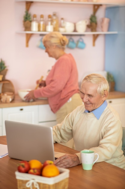 Älterer Mann, der an einem Laptop arbeitet, während seine Frau kocht