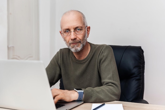 Älterer Mann, der an seinem Laptop arbeitet und in seine Kamera schaut