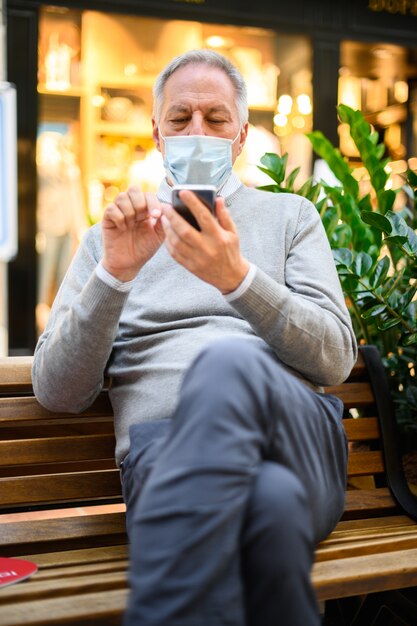 Älterer Mann, der auf einer Bank sitzt und ein Smartphone in einem Einkaufszentrum trägt, das Maske trägt