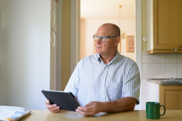 Älterer Mann, der beim Verwenden des digitalen Tablets durch das Fenster denkt