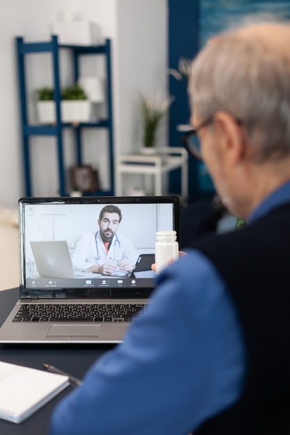 Älterer Mann, der dem Arzt eine Pillenflasche zeigt, während er einen Laptop für die Telemedizin verwendet. Ein älterer Mann, der im Zuge eines Ferngesprächs mit einem Arzt diskutiert, und seine Frau liest ein Buch auf dem Sofa.