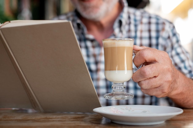 Älterer mann, der draußen an einem cafétisch sitzt und einen kaffee und milch dring hält, während er ein buch liest kaukasischer älterer mann entspannte sich im ruhestand oder urlaub