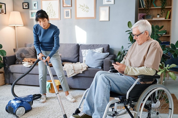 Älterer Mann, der Handy benutzt, während Haushälterin ihm hilft, das Zimmer mit Staubsauger zu reinigen