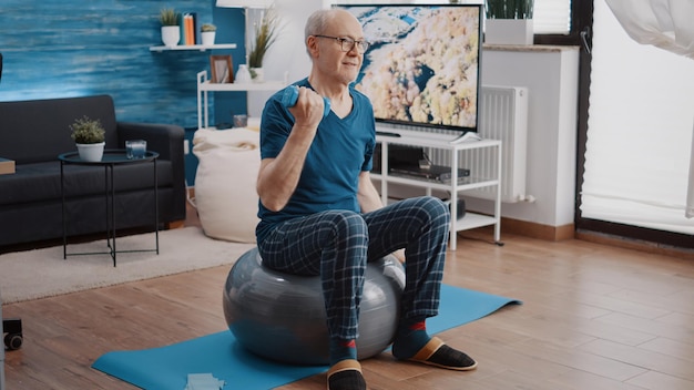 Älterer Mann, der Hanteln hebt und auf einem Fitnessball sitzt, um körperliches Training und Bewegung zu machen. Rentner, der Trainingsaktivitäten mit Gewichten durchführt, um die Armmuskulatur zu Hause zu trainieren.