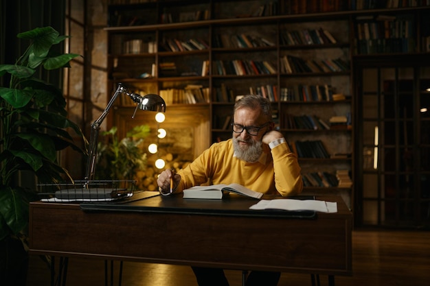 Älterer Mann, der im Heimbüro mit Dokumenten arbeitet. Älterer Mann sitzt am Tisch in der Bibliothek und blickt in die Kamera