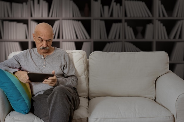 Älterer Mann, der in einem digitalen Tablet liest, das auf dem Sofa im Wohnzimmer des Hauses sitzt