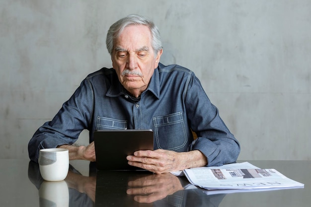 Älterer Mann, der Social Media auf Tablet mit einer Tasse und einer Zeitung auf dem Tisch nutzt
