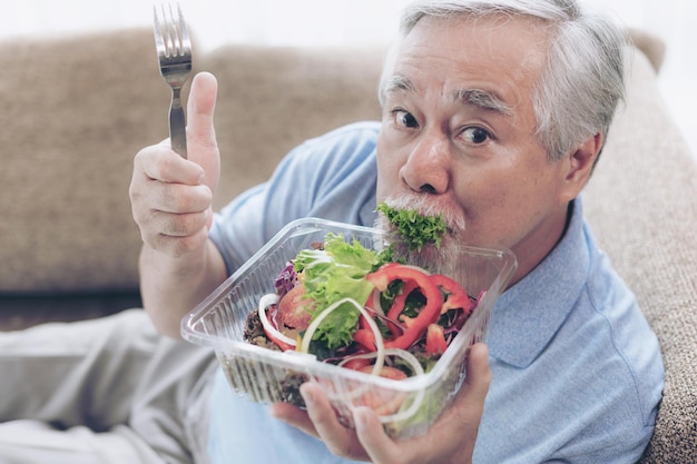 Älterer Mann des Lebensstils fühlen sich glücklich, frischen Salat der Diätlebensmittel auf Sofa zu essen