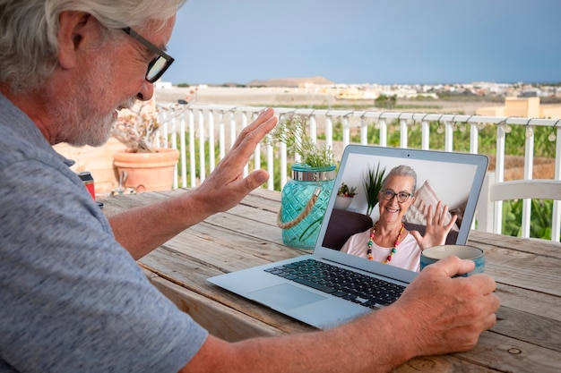 Älterer Mann im Freien auf der Terrasse am Holztisch sitzend mit Laptop im Video-Chat