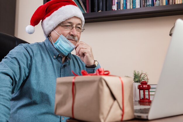 Älterer Mann im Weihnachtsmannhut spricht unter Verwendung des Laptops für Videoanruffreunde und -kinder. Das Zimmer ist festlich eingerichtet. Weihnachten während des Coronavirus.