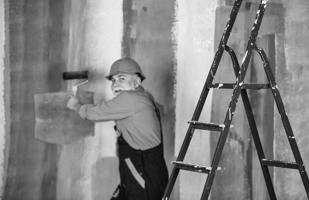 Älterer Mann, Maler, verwendet eine Rolle auf der Leiter, um die Wand zu streichen. Bunter professioneller Maler in Arbeitskleidung