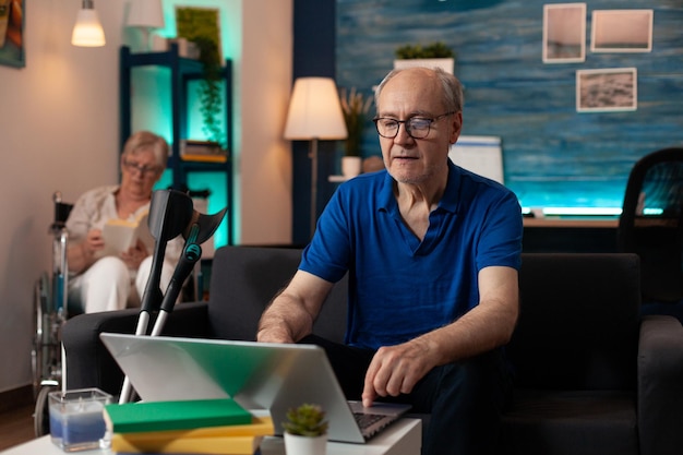 Älterer Mann mit Laptop auf dem Tisch im Wohnzimmer. Rentner mit digitalem Gerät und Krücken auf der Couch, während ältere Frau im Rollstuhl im Hintergrund zu Hause sitzt.