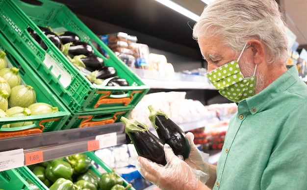 Älterer Mann mit medizinischer Maske und Schutzhandschuhen beim Einkaufen im Supermarkt bei der Auswahl von Gemüse