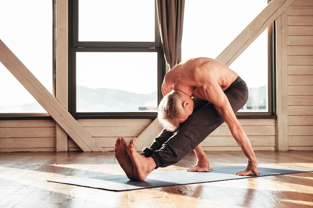 Älterer Mann mit nacktem Oberkörper, der im Studio vor einem Fenster Yoga-klassisches Asana-Training praktiziert