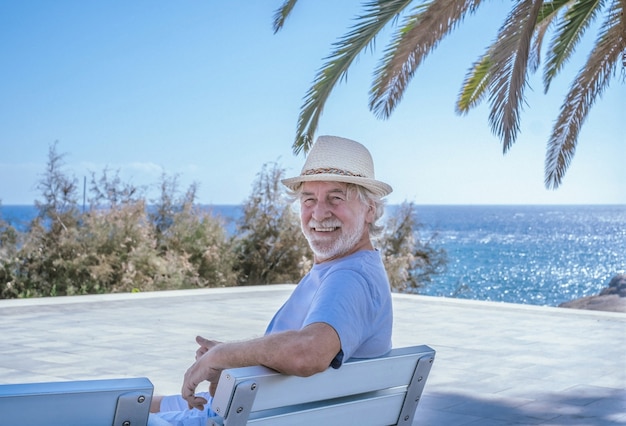 Älterer Mann mit Strohhut, der draußen auf dem Meer sitzt und lächelnd in die Kamera schaut - Horizont über Wasser